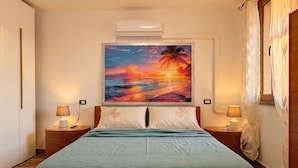 Een slaapkamer met een bed, twee nachtkastjes met lampen, een wandairconditioner en een groot schilderij van een zonsondergang boven de oceaan.