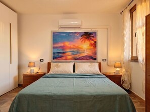 A bedroom with a bed, two bedside tables with lamps, a wall-mounted air conditioner, and a large painting of a sunset over the ocean. - Vacation home Mamau by Interhome (Castelsardo)
