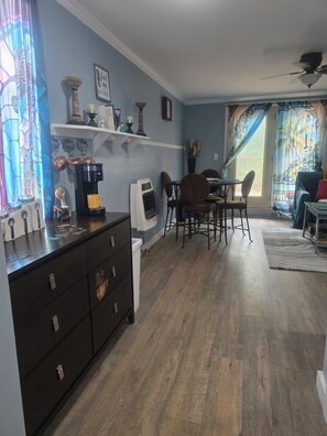 A living room with a coffee maker, a dining table with chairs, a sofa, and a window with curtains.