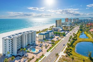 A beachfront hotel with multiple buildings, a swimming pool, and a road with palm trees. - Beachfront 2B/2B Vacation Condo at Shamron Beach! Pool! Book Now For Dream Vacation! (Fort Myers Beach)