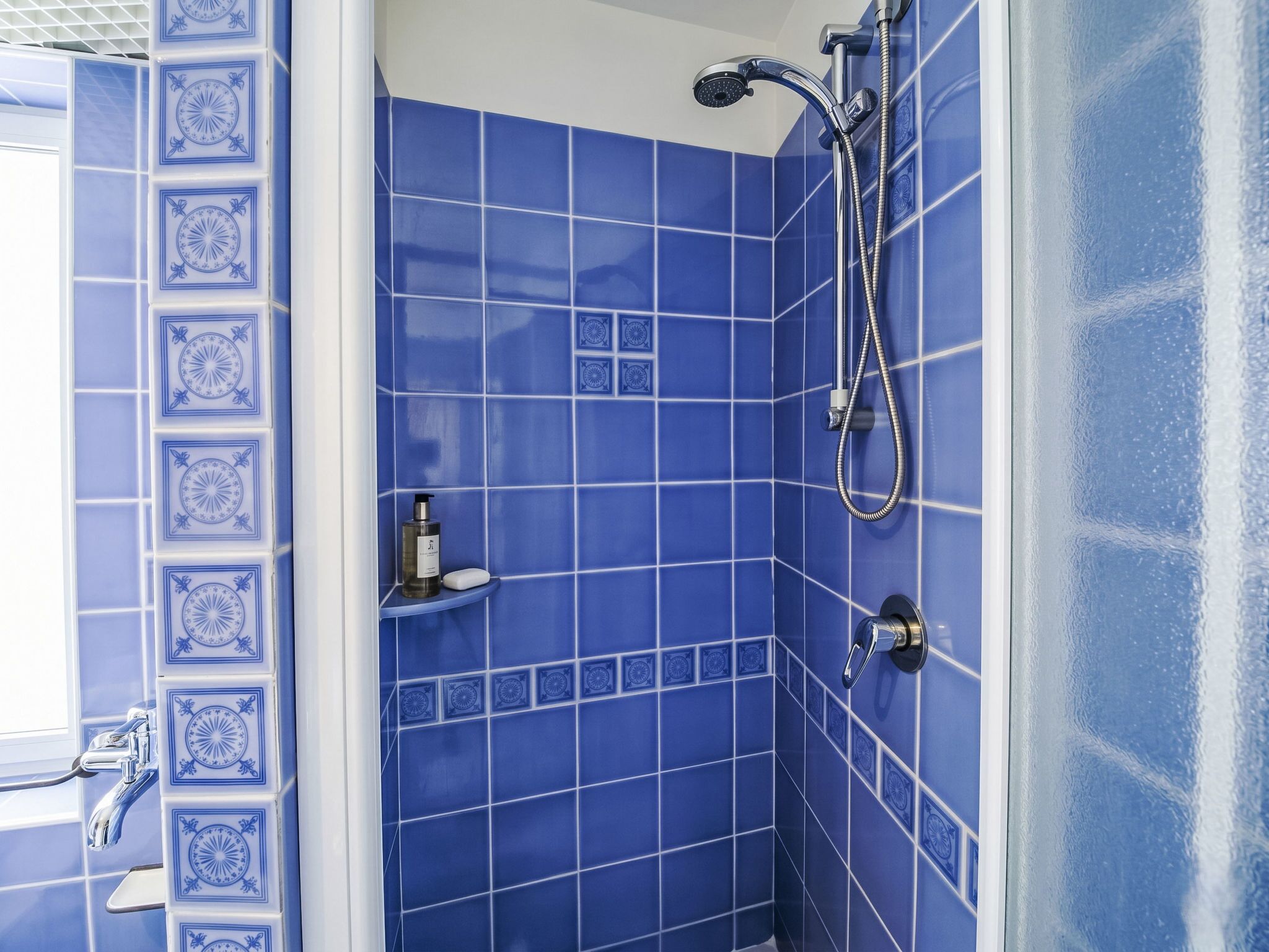 A bathroom with blue tiles, a shower, and a shelf with toiletries.