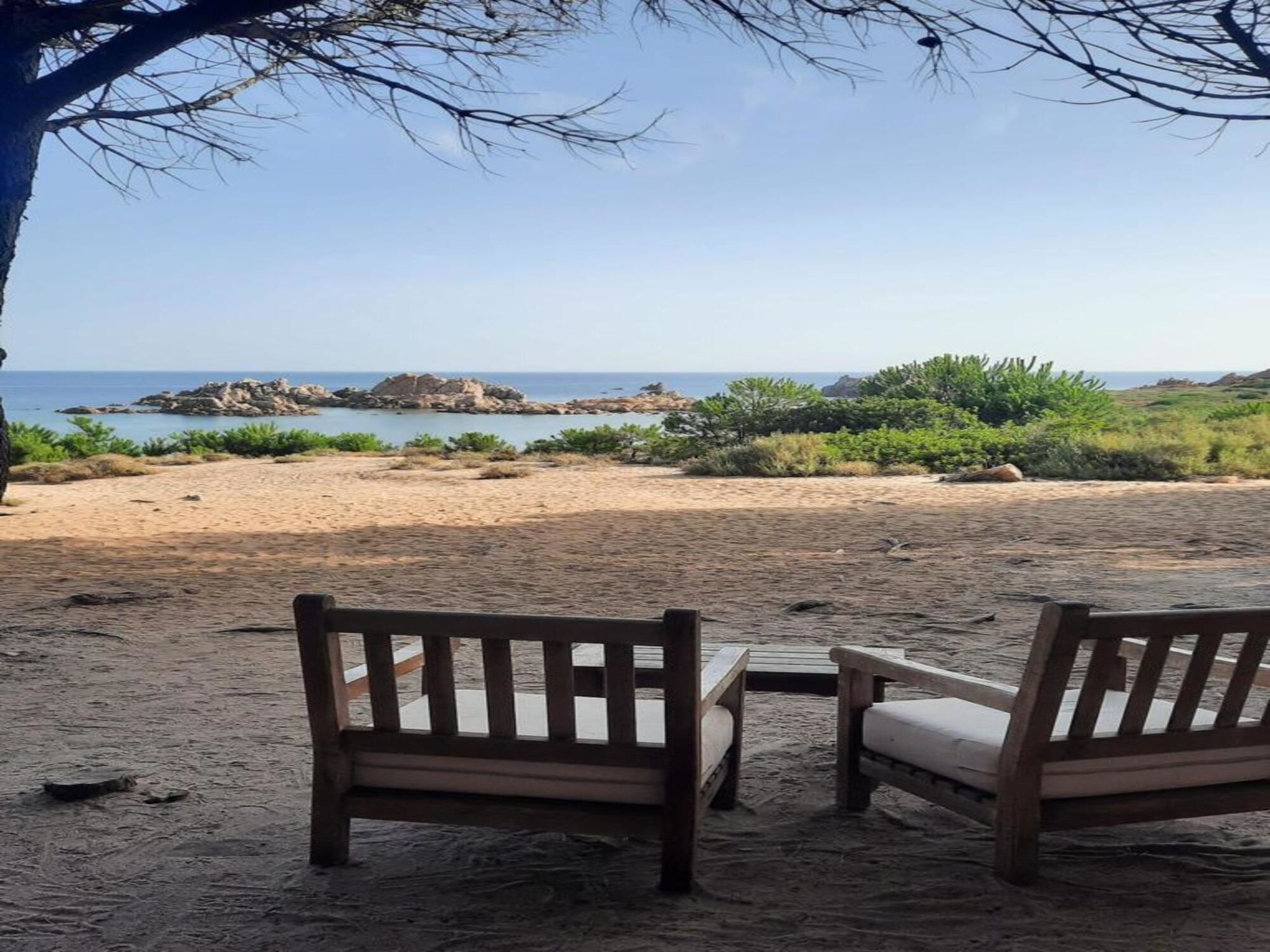 A beach with two wooden benches facing the sea, a clear sky, and rocky outcrops in the distance.