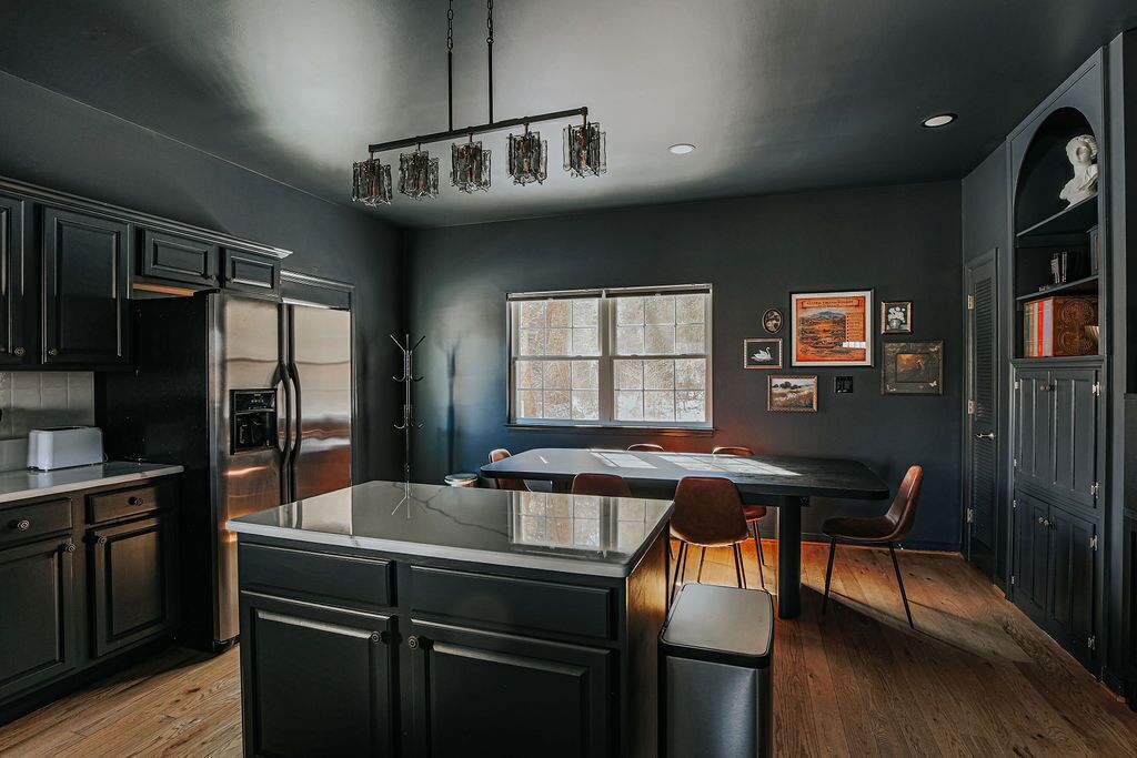 A modern kitchen with dark cabinetry, a stainless steel refrigerator, and a central island. There is a dining area with a table and chairs, and a large window providing natural light.