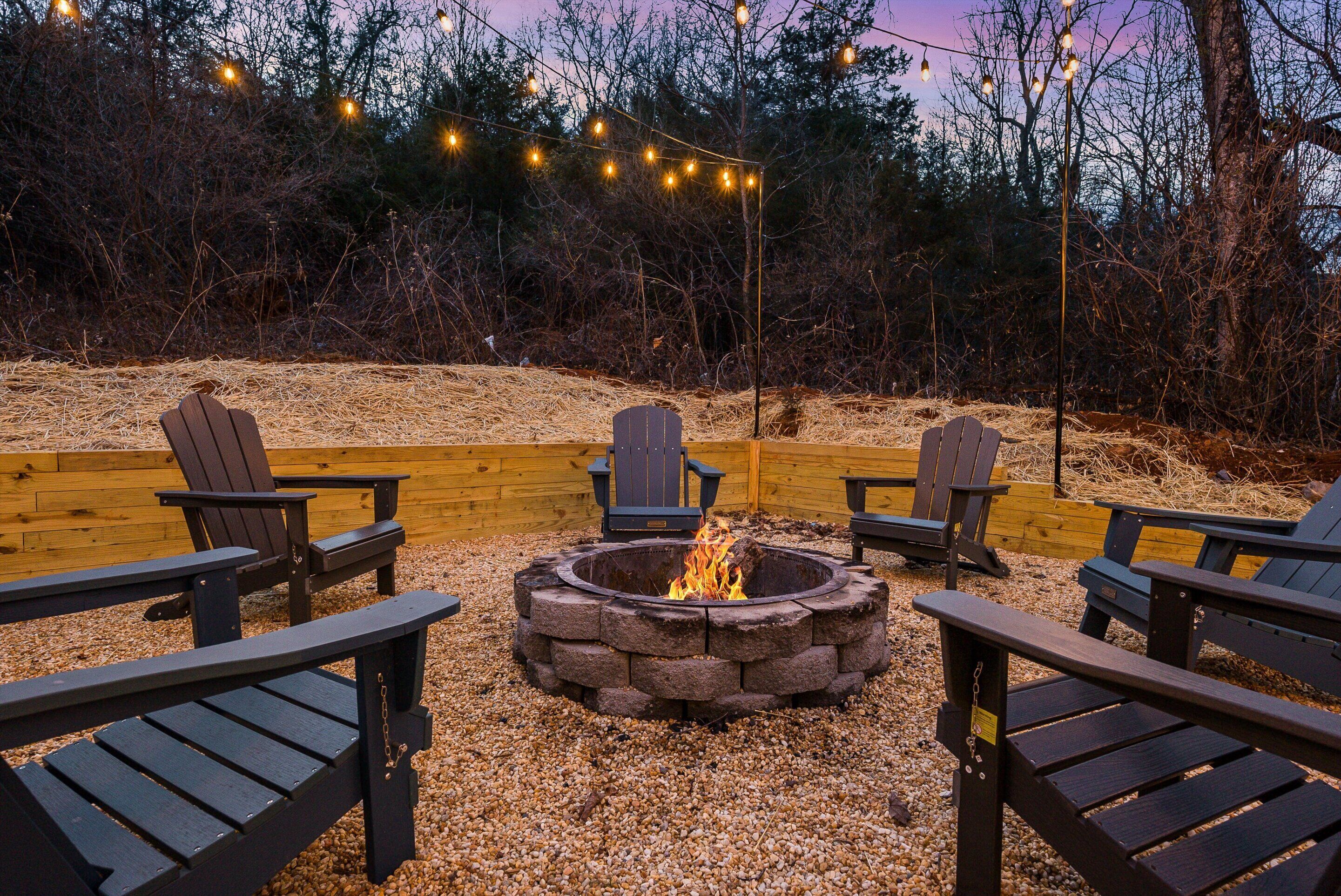 A fire pit surrounded by Adirondack chairs, with string lights overhead and a wooden fence in the background.