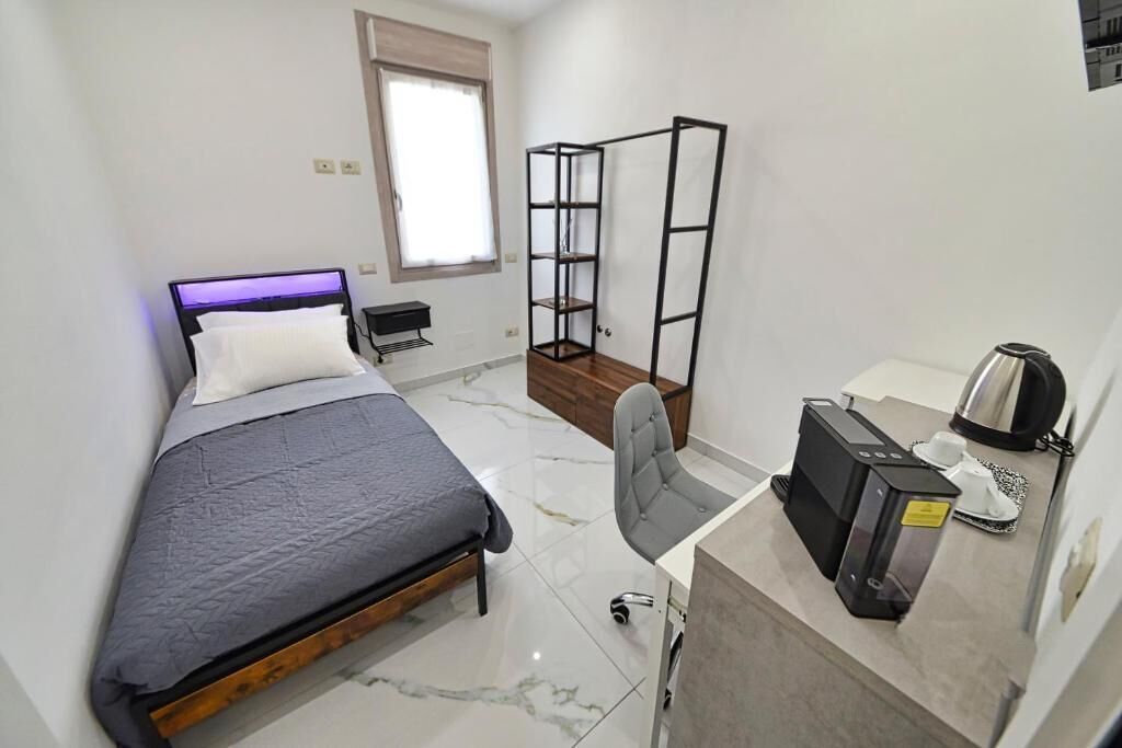 Ein kleines Hotelzimmer mit einem Bett, einem Schreibtisch mit Kaffeemaschine, einem Sessel und einem Bücherregal.