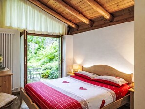 Camera da letto con soffitto in legno, un letto con biancheria rossa e bianca, un comodino con una lampada e vista sul verde all'esterno.