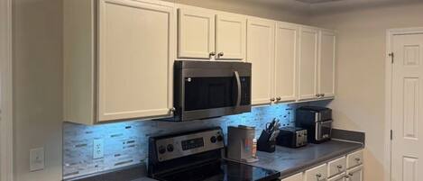 A kitchen with white cabinets, a black microwave, a stove, a trash can, and a ceiling light fixture.