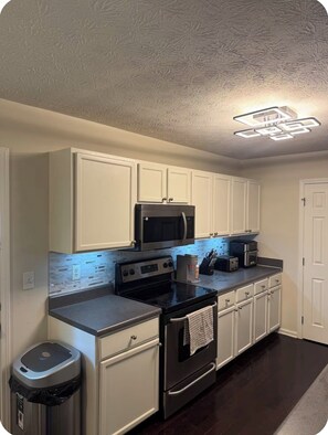 A kitchen with white cabinets, a black microwave, a stove, a trash can, and a ceiling light fixture.
