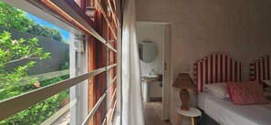 Un dormitorio con cama, mesita de noche, lámpara, espejo y vistas al exterior con vegetación.