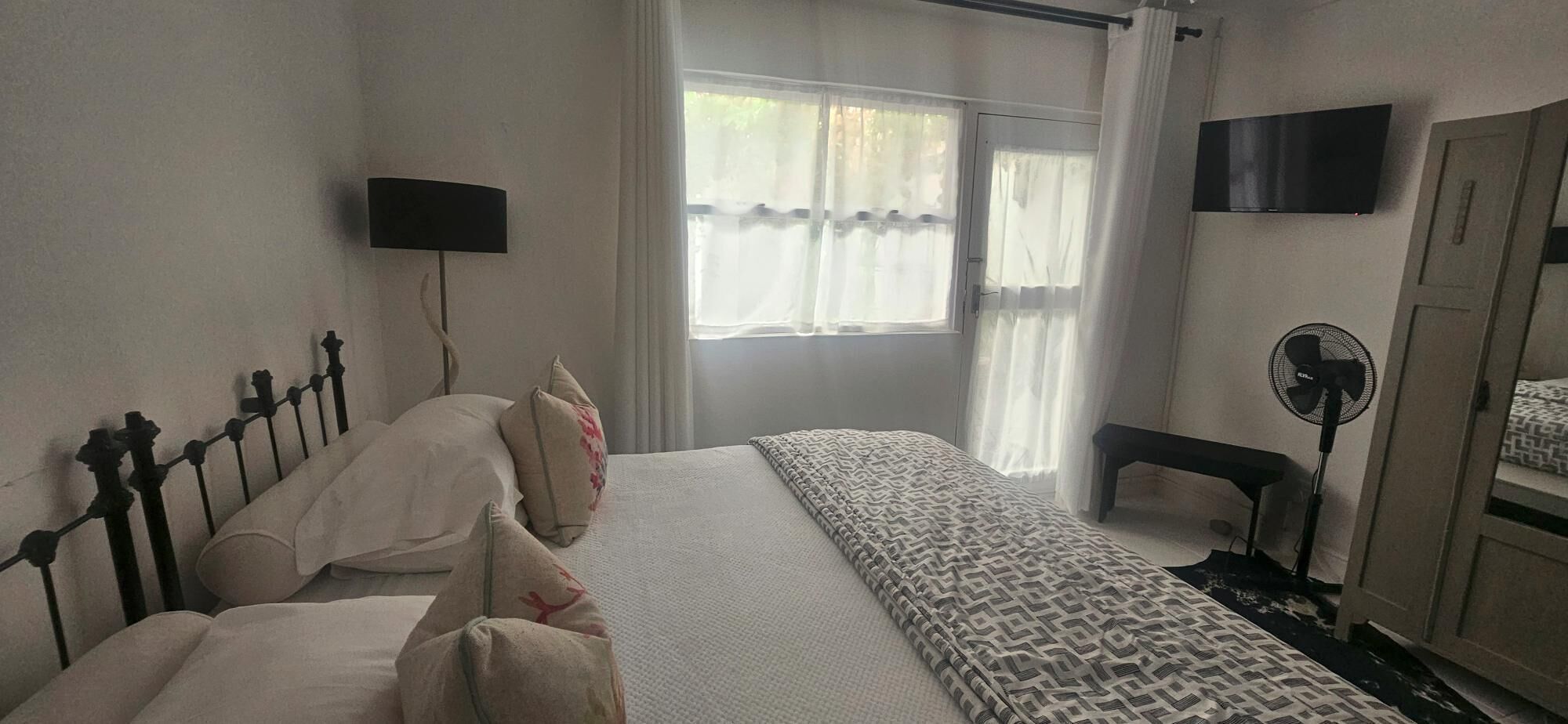 A bedroom with a bed, a bedside table, a chair, a standing fan, and a TV mounted on the wall.