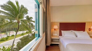 A hotel room with a bed, a nightstand, a lamp, a window with a view of palm trees and a road. - Viona Beach Hotel - Marina Phu Quoc (Phu Quoc)