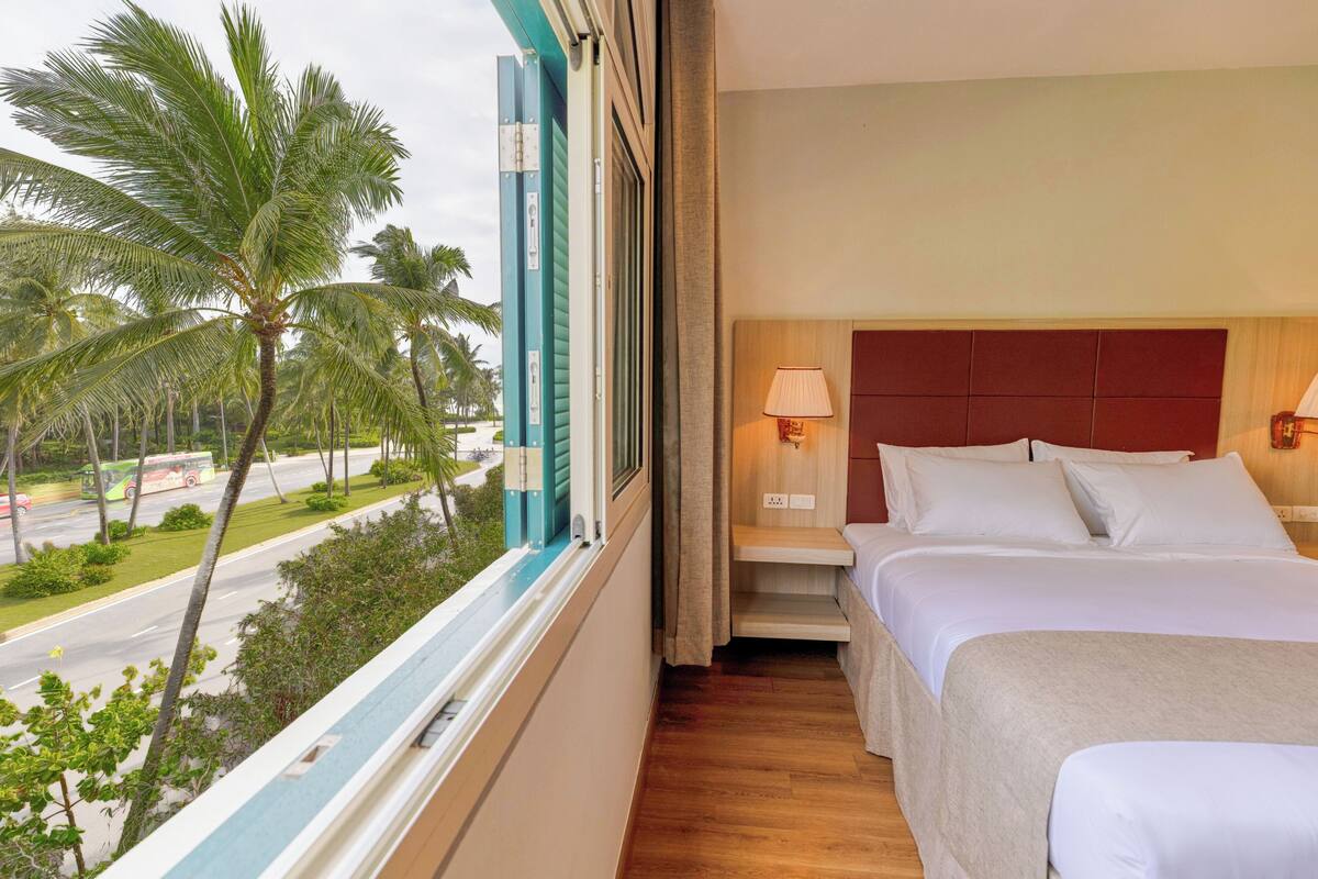 Habitación de hotel con cama, mesita de noche, lámpara, ventana y vista a palmeras y una carretera.