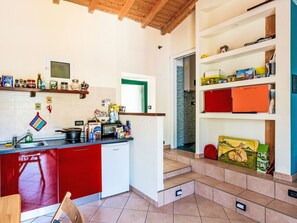 Eine Küche mit roter Arbeitsplatte, einem Spülbecken und verschiedenen Küchenutensilien. Ein Fenster mit grünem Rahmen und eine Tür zu einem anderen Raum sind ebenfalls vorhanden.