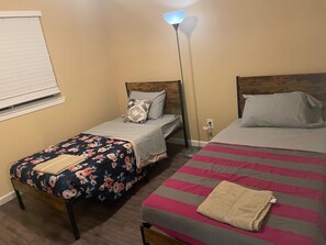 Ein Zimmer mit zwei Betten, eines mit Blumenmuster und das andere mit Streifenmuster, einer Stehlampe und einem Fenster mit Jalousien.
