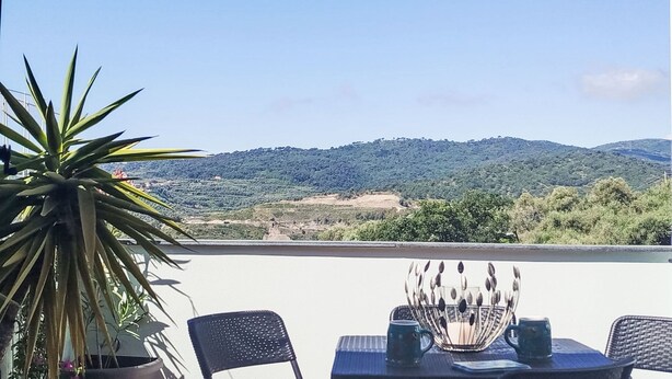 En terrasse med et bord, stole, en potteplante og udsigt over et bakket landskab.