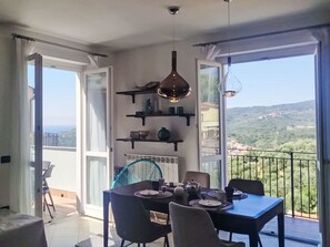 A dining area with a table set for four, a blue chair, and a view of a green landscape through large windows. - Apartment Melba by Interhome (Civezza)