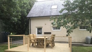 Eine Holzterrasse mit Tisch und Stühlen, ein Haus mit grauem Dach und ein Baum im Hintergrund.