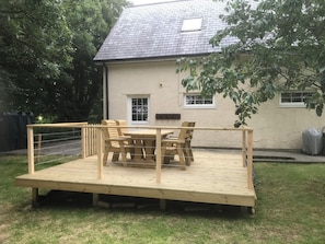 A wooden deck with a table and chairs, a house with a grey roof, and a tree in the background. - Swallows Cottage (Saint Giles on the Heath)