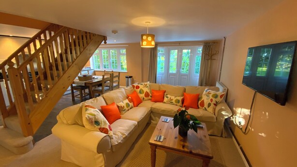 Ein Wohnzimmer mit einer Holztreppe, einem Sofa mit orangefarbenen Kissen, einem Couchtisch und einem an der Wand montierten Fernseher.