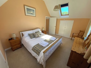 A bedroom with a large bed, a nightstand, a chair, a wardrobe, and a window with a view. - Swallows Cottage (Saint Giles on the Heath)
