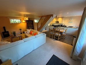 A cozy living room with a sofa, a dining area with chairs, a staircase, a kitchen with cabinets, and a bookshelf. - Swallows Cottage (Saint Giles on the Heath)