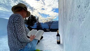 Uma pessoa lendo um livro ao ar livre, com uma garrafa e um copo sobre uma superfície branca.