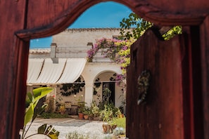 A courtyard with a building featuring arches and a tiled roof, surrounded by potted plants and flowering vines.