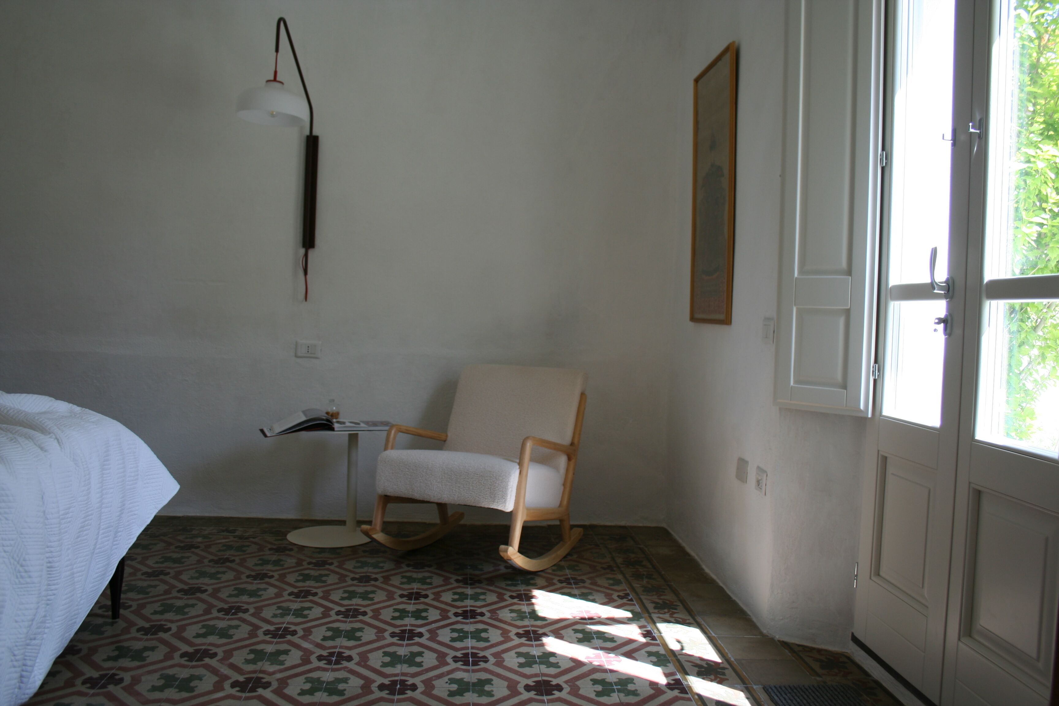A room with a bed, a rocking chair, a small table, a wall-mounted lamp, and a window with a view of greenery.