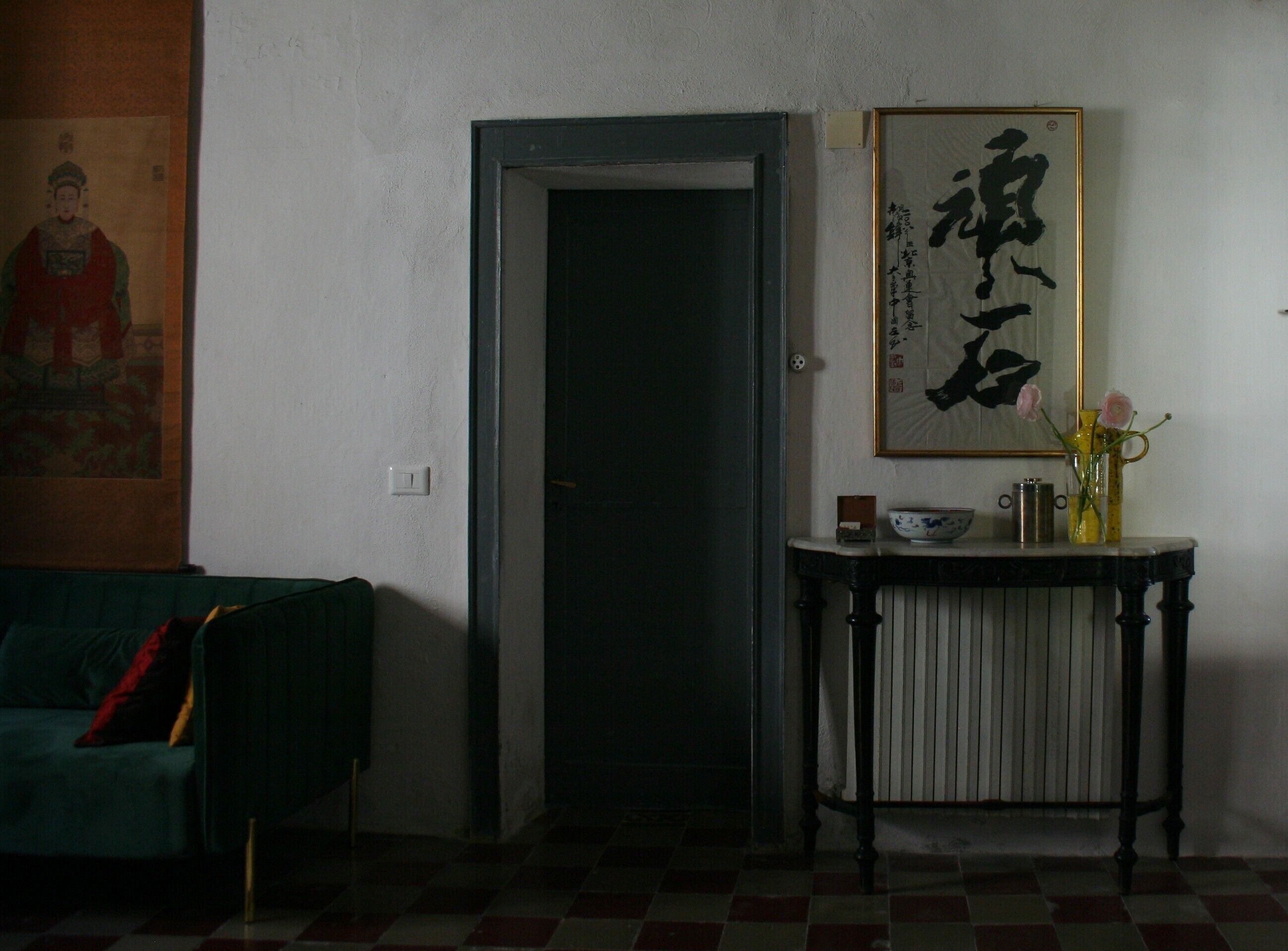 Una habitación con una puerta cerrada, un cuadro caligráfico enmarco, una pequeña mesa con un florero y un cojín sobre un sofá verde.
