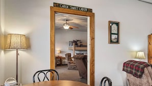 A room with a wooden door labeled 'Cabin Sweet Cabin,' a dining table with chairs, a lamp, a clock, and a cozy seating area with a chair and a blanket. - The Goose Cabin | 5-Cabin Compound, Sleeps 30 (Decatur)