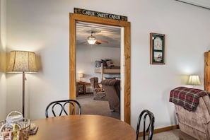 Una habitación con puerta de madera que dice 'Cabin Sweet Cabin', una mesa de comedor con sillas, una l ámpara, un reloj y un rincón acogedor con una silla y una manta.