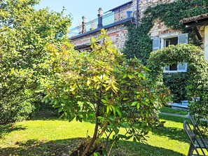 Ein Garten mit verschiedenen Bäumen und Sträuchern, ein Steinhaus mit Balkon und einem klaren, blauen Himmel.