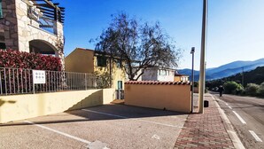 Un'area di parcheggio con superficie pavimentata, un marciapiede in mattoni e un edificio con facciata in pietra e recinzione in metallo.