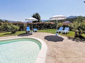 Ein Schwimmbad mit Liegestühlen und Sonnenschirmen, umgeben von Grünflächen und Hügeln.