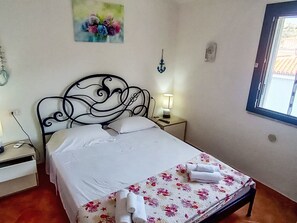 Ein ordentlich bezogenes Bett mit weißen Leinen, einer decke mit Blumenmuster und zusammengerollten Handtüchern. Ein Nachttisch mit Lampe, eine Wanduhr und ein Blumenbild sind ebenfalls vorhanden.