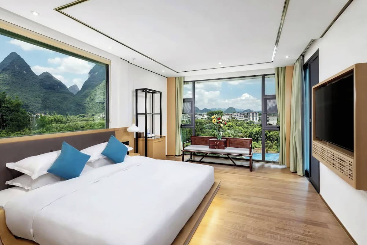 Una habitación de hotel moderna con una cama grande, televisión, balcón con vistas a montañas y escritorio con lámpara.