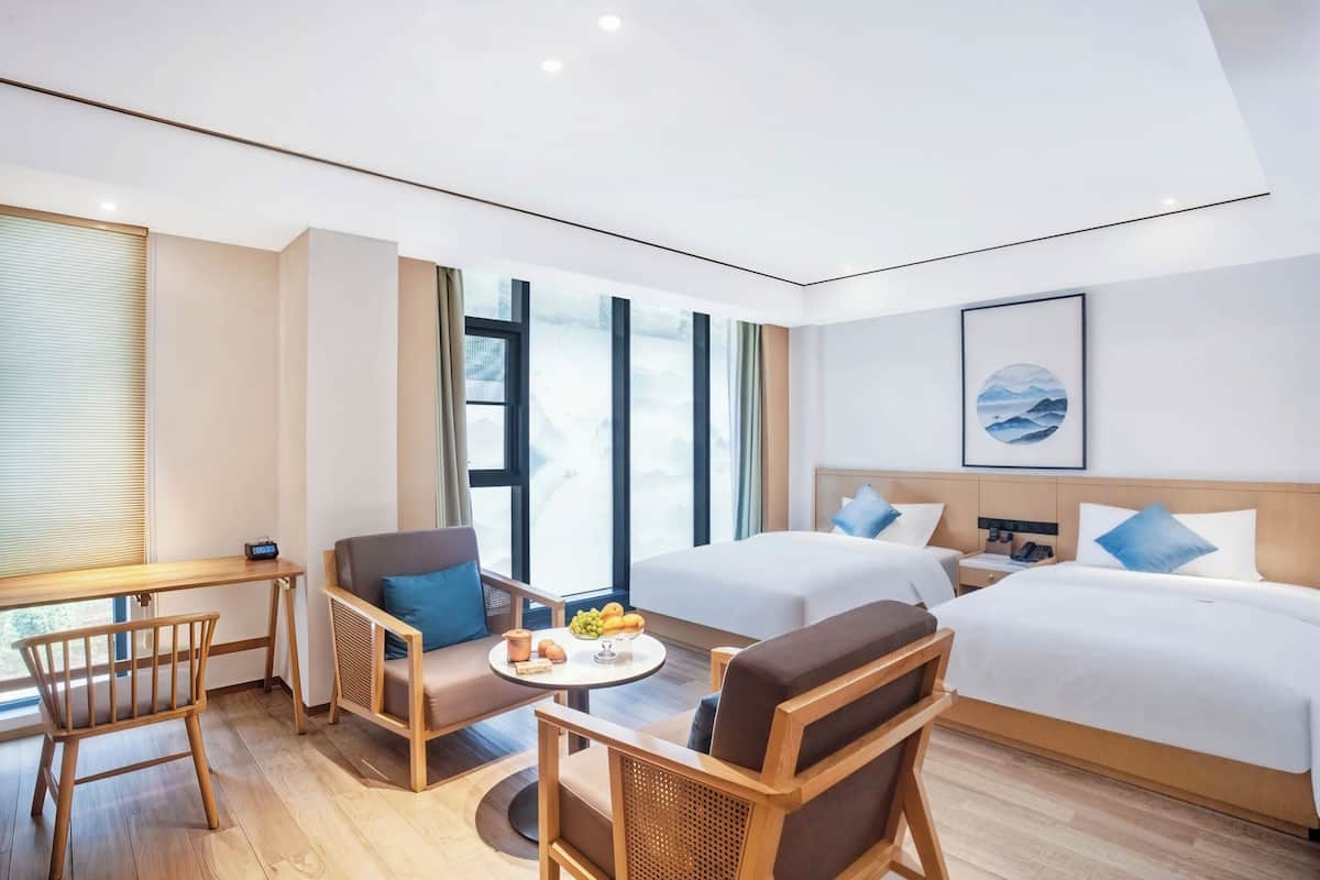 Habitación de hotel con dos camas, un escritorio, una silla, una mesita con frutas y un ventanal grande.