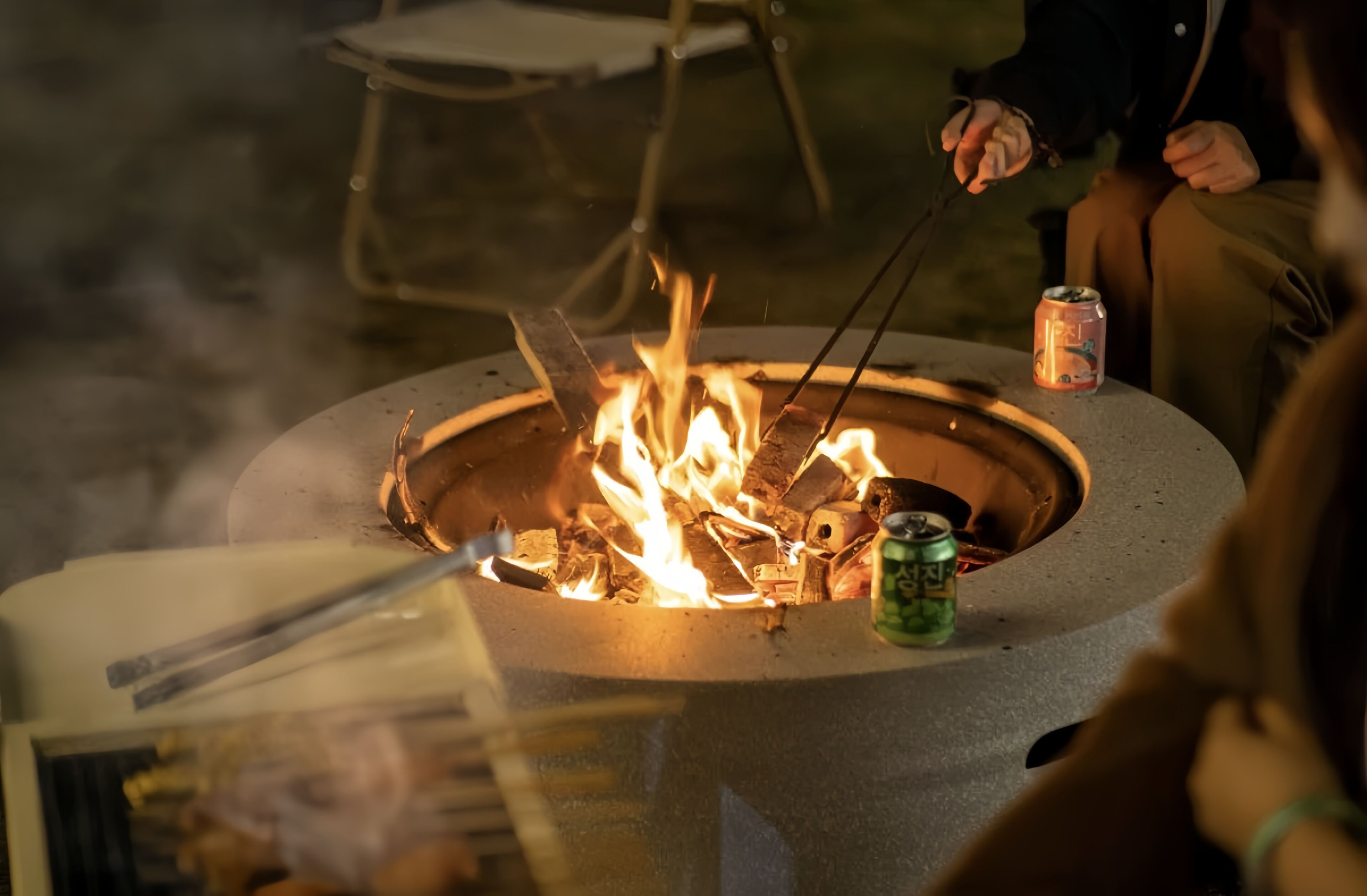 A fire pit with flames, a can of beer, and a person holding a stick.