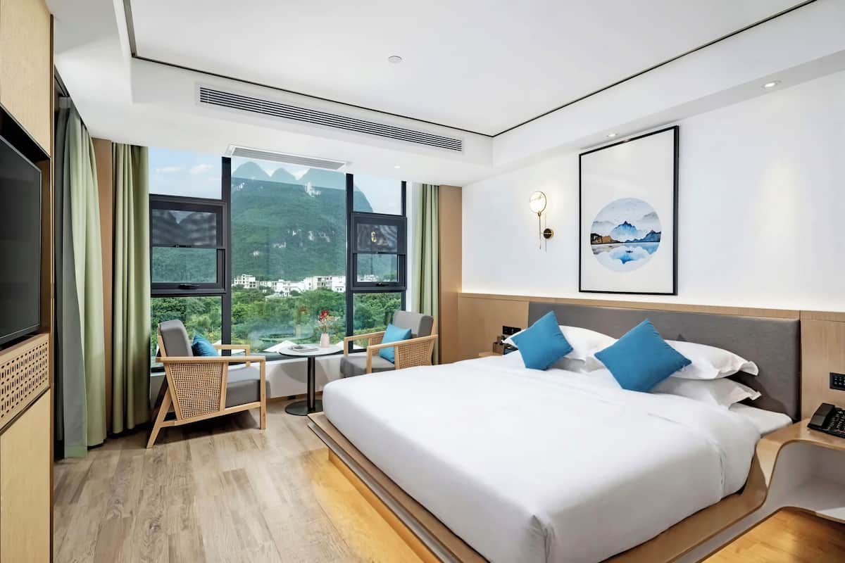 Una habitación de hotel moderna con una cama grande, televisión, escritorio con silla y vistas a montañas a través de amplios ventanales.