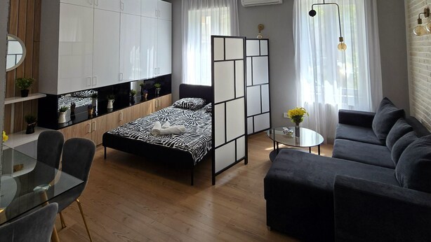 En moderne stue med sofa, spisebord med stole, seng med sort-hvidt sengetæppe, og et køkken med skabe og bordplade.