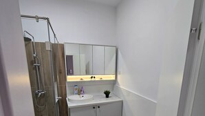 Et moderne badeværelse med en brusekabine i glas, en hvid vask, et spejl over vasken og en dør, der fører til et andet rum.