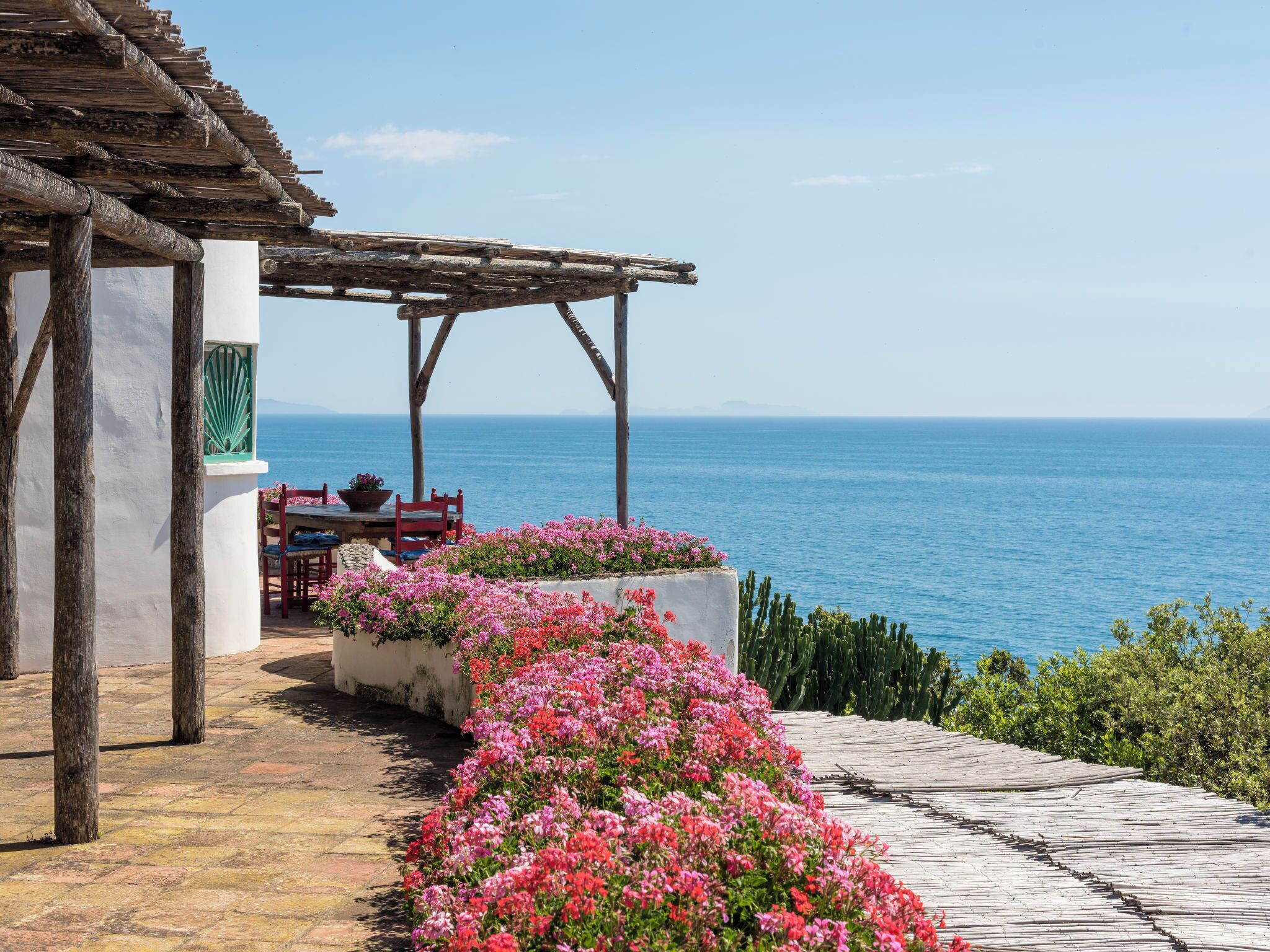 A seaside patio with a wooden pergola, a table with chairs, and a flower bed.