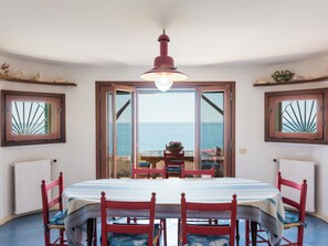 A dining area with a table and chairs, a view of the sea through a door, and a hanging light fixture.