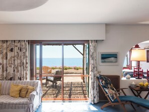 A room with a sofa, a chair, a table with a lamp, a painting, and a view of the sea through a glass door.