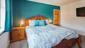 Een slaapkamer met een groot bed, een houten hoofdeinde, een nachtkastje, een aan de muur bevestigde televisie en een raam met gordijnen.
