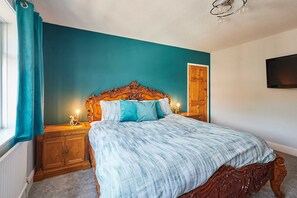 A bedroom with a large bed, wooden headboard, teal walls, a wooden nightstand, a wall-mounted TV, and a window with curtains. - Host Stay Roseberry Avenue (Skegness)