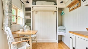 A cozy room with a wooden table, a chair, a bed with a white headboard, and a shelf with decorative items. - Host Stay Shepherds Delight (Harleston)