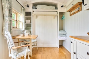 A cozy room with a wooden table, a chair, a bed with a white headboard, a shelf with decorative items, and a window with curtains. - Host Stay Shepherds Delight (Harleston)