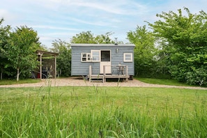 A small, single-story cabin with a porch, surrounded by a grassy field and trees. - Host Stay Shepherds Delight (Harleston)