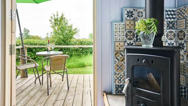 Una terraza de madera con sillas de mimbre y una mesa, un paraguas verde y un estufa de leña negra con azulejos decorativos.